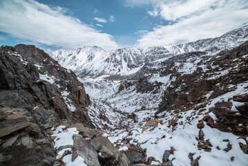 Descent along thin rocky gorge down into snow-covered alpine valley. Landscape from above to snowy stony couloir. White snow on stones in high mountains. Top view to snow-white steep slope among rocks