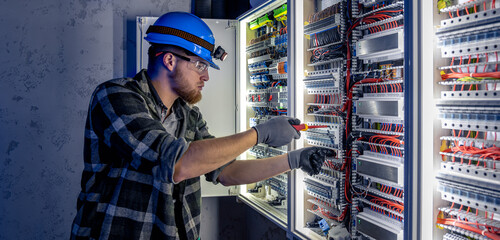 A male electrician works in a switchboard in overalls against the backdrop of emergency lighting.