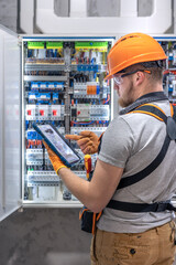 A male electrician in overalls, focused on work in switchboard with fuses, using tablet.