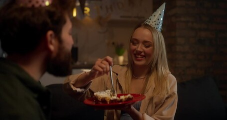 Happy blonde girl in a party cap feeds her boyfriend cake with a fork during a birthday party at home in the evening