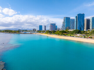 Naklejka premium Aerial view of Honolulu, Oahu, Hawaii, showing turquoise waters, sandy beach, greenery, a road with vehicles, and modern high rise buildings under a blue sky.