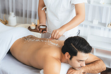 Blissful man customer having exfoliation treatment in luxury spa salon with warmth candle light ambient. Salt scrub beauty treatment in health spa body scrub. Quiescent