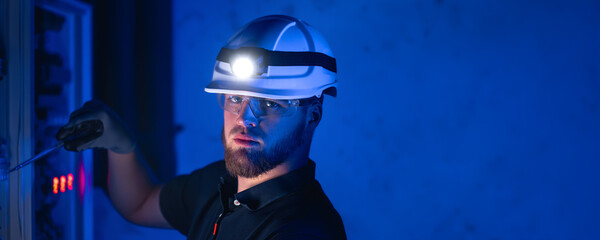 A male electrician works in a switchboard in overalls against the backdrop of emergency lighting.