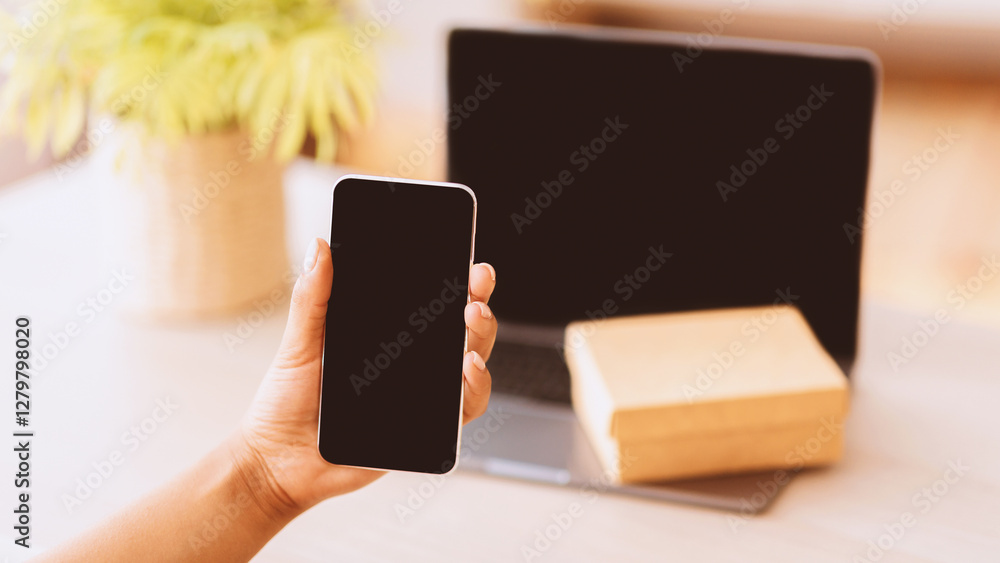 Poster Technology Concept. Unrecognizable afro lady holding and showing smartphone with black blank screen for mock up. Laptop computer with present box or delivery package on keyboard in blurred background