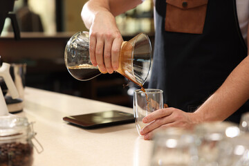 Barista pouring coffee from glass coffeemaker into cup at table in cafe, closeup