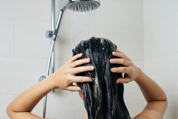 Naklejka premium A person with wet, dark hair lathers shampoo in the shower, water cascading from the overhead showerhead.