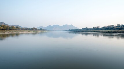 Fototapeta premium Calm river flows through a peaceful valley with distant mountains under a hazy sky during early morning.