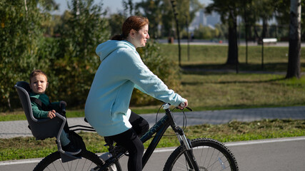 Fototapeta premium Caucasian woman riding a bicycle with her toddler son sitting behind her in a child seat. 