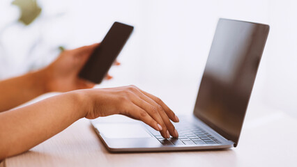 Busy Worker. Side View Of Young Unrecognizable African American Lady Holding Mobile Phone With Blank Empty Screen, Sitting At Table With Computer And Typing On Laptop Keyboard, Blurred Background