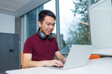 Fototapeta premium A young Asian male college student focused and happily doing work in a classroom on his laptop. He has a headphone around his neck and is wearing a typical university student outfit