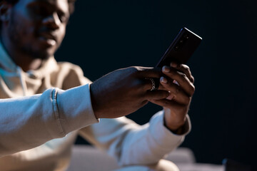 Man at home in dark living room writing messages on phone. BIPOC person holding cellphone, enjoying...