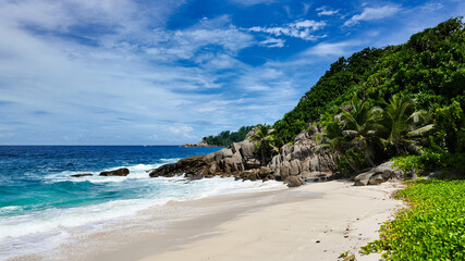 Sandy shoreline with crashing turquoise waves, lush greenery, and tropical plants under a bright blue sky. Seychelles, Mahe. Anse Boileau.