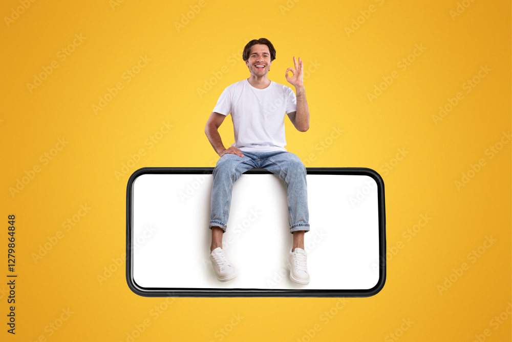 Poster cheerful young man sitting on big huge horizontal new cellphone with white empty screen and showing 