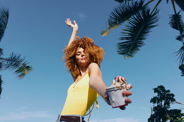 Joyful woman dancing outdoors under palm trees in a bright yellow top with a drink in hand, set...