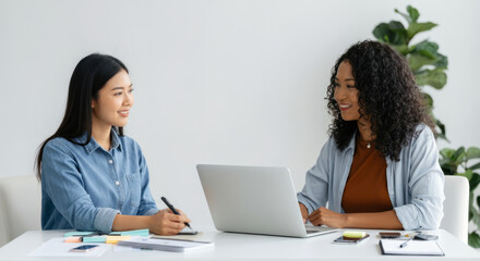 Two women are sitting at a table, one of them is writing on a laptop