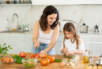 Cheerful middle-aged woman and her daughter making vegetable salad in the kitchen
