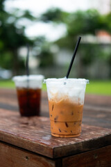 A glass of iced Thai milk tea and black coffee background in the plastic cup after drink finishes on wood table at garden cafe