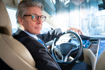 A senior gentleman with glasses and grey hair is seated in a sleek, contemporary luxury e-car,...