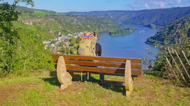 Backpacking hiker relaxing on wooden bench with panoramic view of the surrounding Rhine River Valley and the village of Bacharach on a sunny summer day, Germany