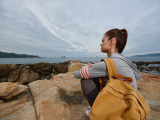 Young woman contemplating by the sea, wearing casual outdoor clothing and a stylish backpack,...