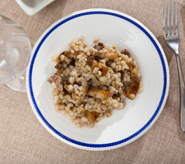 on red tablecloth with golden stars is terracotta-colored plate with portion of stewed barley porridge with fried lard,complemented by glass of red wine and pickled cucumbers