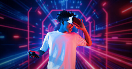 Fototapeta premium Teenager holding joystick and wearing VR goggles while enjoy playing games at neon abstract background. African man enjoy controlling controller and using augmented reality technology. Ingenuity.
