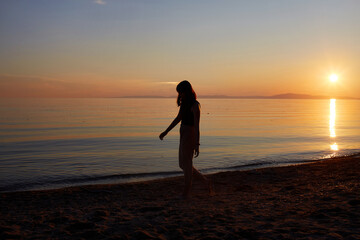Fototapeta premium A beautiful girl walks along the sandy beach at sunset. Summer travel, seaside holidays.