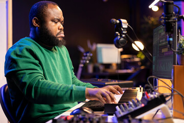 African american audio technician using piano synthesizer keys to add audio effects in post production, editing tunes with digital software and mixing console. Male artist in home studio.