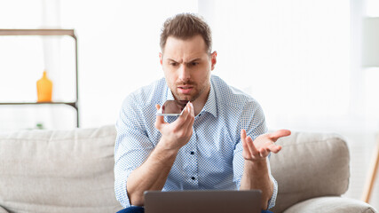 Confused adult businessman talking on phone, holding cellphone near mouth and gesturing, looking at laptop