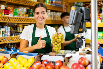 Smiling female shopping assistant weighing grapes in grocery shop, showing thumbs up