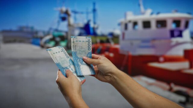 Hands holding chilean pesos outdoors in a seaside port with boats, depicting business and investment activities in a maritime city setting.