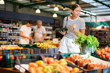 Positive young female shopper making purchases in supermarket, looking with interest for ripe organic courgettes