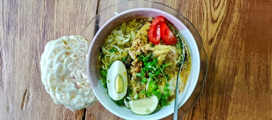 A bowl of traditional Indonesian Soto with boiled egg, crackers, tomatoes, lime, and shredded chicken on a wooden table. A flavorful soup dish.  
