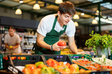 Confident young guy, grocery store worker, preparing fresh vegetables for sale, arranging ripe red...