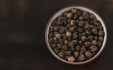Close-up of black peppercorns in a ceramic bowl on a dark wooden background. Aromatic spice used in cooking, perfect for food photography and culinary designs.
