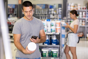Young adult man scanning barcode on can with paint using smartphone while buying materials for...
