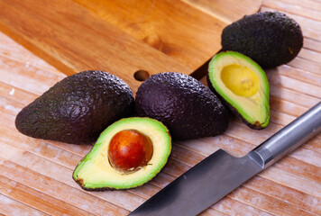 Whole and sliced avocados on wooden board with kitchen knife.