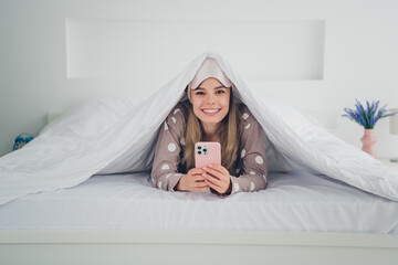 Obraz premium Joyful young woman in pajamas using smartphone under cozy blanket at home during daylight
