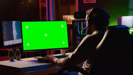 Man in dimly lit apartment playing video games on green screen gaming PC at computer desk, enjoying day off from work. Gamer battling enemies in online multiplayer shooter on mockup monitor © DC Studio