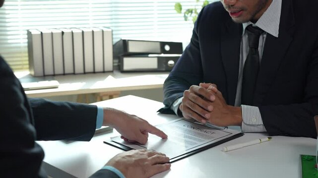 Business partners discussing terms of contract, pointing at specific clauses, gesturing, and finally shaking hands after reaching an agreement in a bright office environment