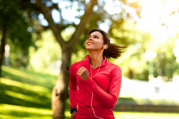 Fototapeta premium Joyful black girl jogging by summer park, listening to music, enjoying fresh air, copy space