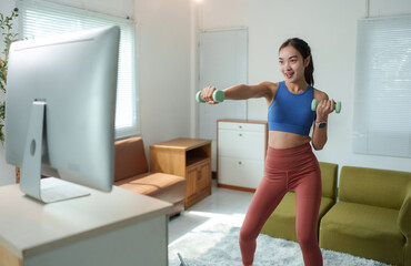 Young woman exercising with dumbbells in a bright living room, following a virtual workout on a computer screen. She is focused and energetic, maintaining a healthy lifestyle