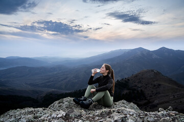 Hydration break in the mountains. Woman drinking water while sitting on a cliff's edge during a hike. Staying hydrated while hiking, trekking, exploring nature, active lifestyles, hydration awareness.