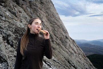 Friendly woman hiker eating granola muesli bar standing in the mountains. Female traveler eating a...