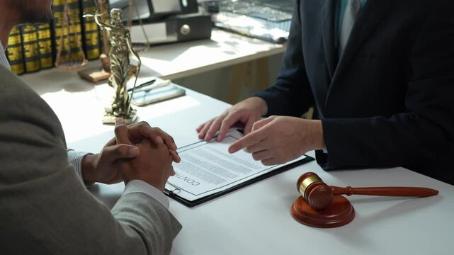Professional lawyer pointing at specific contract details, explaining legal terms to attentive client near judge gavel and themis statue on pristine white table within law firm setting