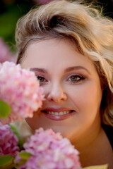 Naklejka premium Portrait of a beautiful fat girl in a blue dress, against the background of a wall of live growing pink gartenias. The plus size woman enjoys the beauty of flowers