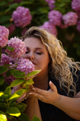 Fototapeta premium Portrait of a beautiful fat girl in a blue dress, against the background of a wall of live growing pink gartenias. The plus size woman enjoys the beauty of flowers