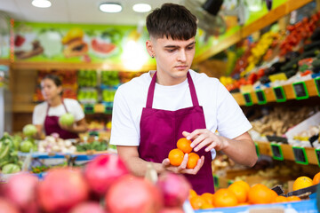 Hardworking young salesman working in a vegetable store puts fresh oranges on the counter for sale