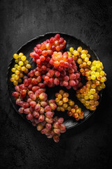 High contrast image of bunches of red and yellow grapes on a black plate against a black textured background with dramatic lighting. Top view.