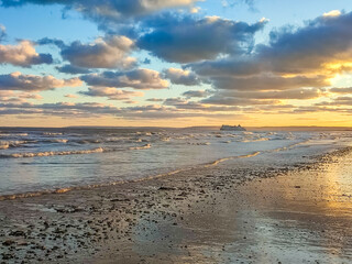 Calm waves lap at the shore as the sun sets behind clouds, creating a serene coastal atmosphere
